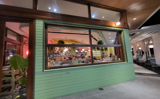 View of Spice Market Thai restaurant from the outside, featuring Smartech's Folding Windows partially open, merging indoor and outdoor spaces. The windows’ wood-grain frames complement the green exterior walls, inviting guests into the warmly lit, bustling interior.