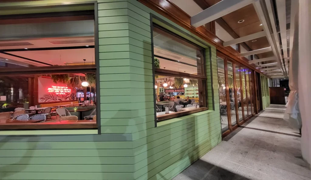 A side view of Spice Market Thai restaurant's dining area through Smartech's Folding Windows, highlighting the seamless integration with the building's green siding and wooden framework. The interior is cozy and vibrant, with diners enjoying their meals.